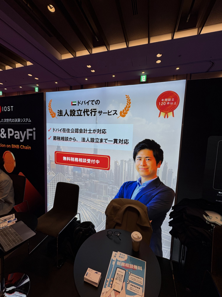Booth display with a smiling man in a blue suit on a large poster, Japanese text and a city skyline background, plus brochures on a round table in front.